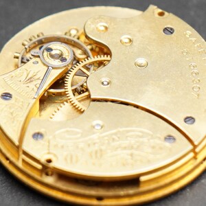 Puede incluir: Primer plano de un movimiento de reloj de bolsillo de oro con engranajes intrincados y una inscripci&oacute;n de barril de seguridad. La esfera del reloj est&aacute; grabada con un dise&ntilde;o floral.