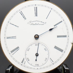 Puede incluir: Esfera de reloj de bolsillo blanca con marcadores de hora en n&uacute;meros romanos negros y agujas negras. La esfera del reloj tiene el texto "American Waltham" impreso.
