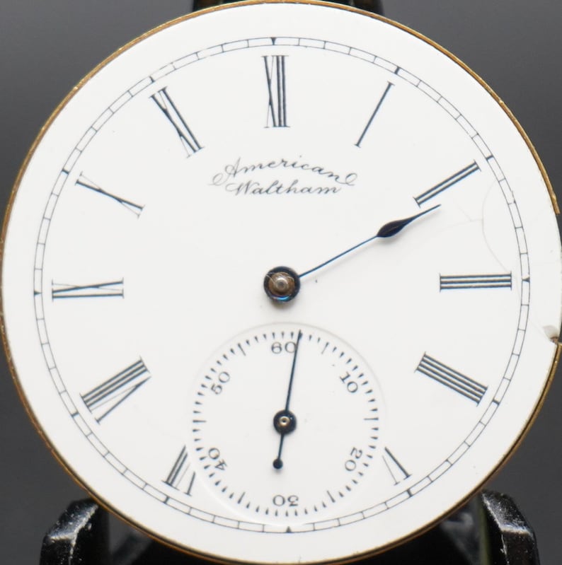 Puede incluir: Esfera de reloj de bolsillo blanca con marcadores de hora en n&uacute;meros romanos negros y agujas negras. La esfera del reloj tiene el texto "American Waltham" impreso.