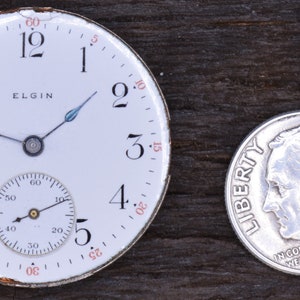 Puede incluir: Un reloj de bolsillo blanco con n&uacute;meros romanos negros y un subdial m&aacute;s peque&ntilde;o. La esfera del reloj dice "ELGIN" y tiene una carcasa plateada. El reloj est&aacute; al lado de una moneda de diez centavos estadounidense de plata con el a&ntilde;o 1964.