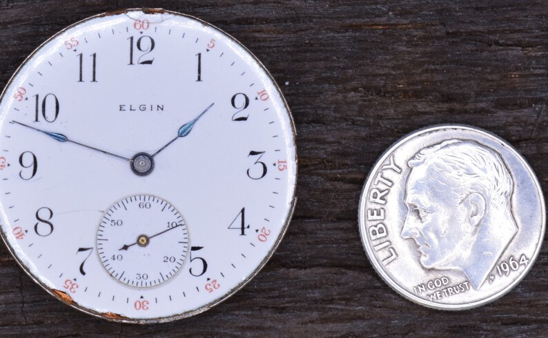 Puede incluir: Un reloj de bolsillo blanco con n&uacute;meros romanos negros y un subdial m&aacute;s peque&ntilde;o. La esfera del reloj dice "ELGIN" y tiene una carcasa plateada. El reloj est&aacute; al lado de una moneda de diez centavos estadounidense de plata con el a&ntilde;o 1964.