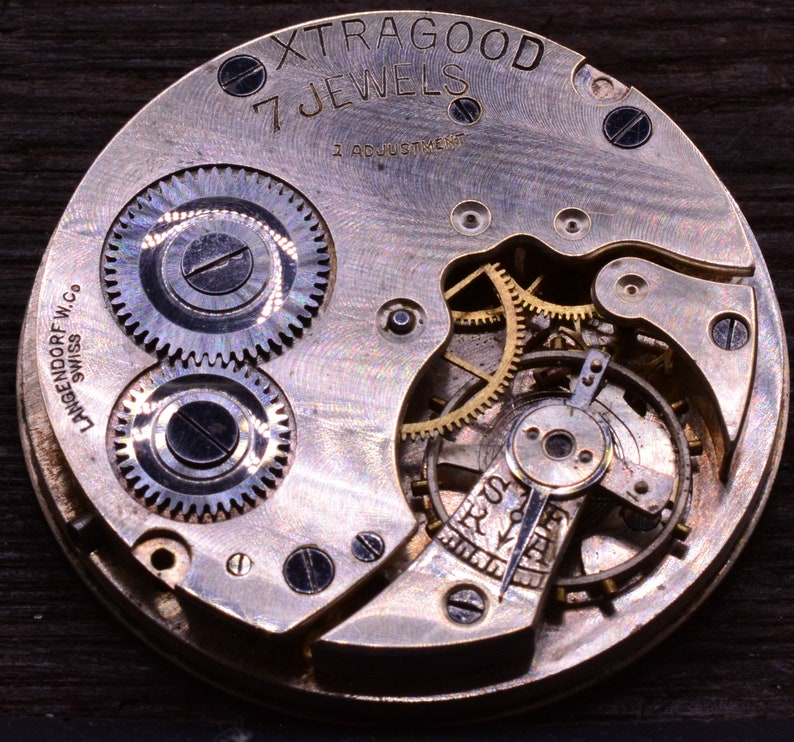 Pu&ograve; includere: Primo piano di un movimento di orologio argentato con il testo "Xtragood 7 Jewels 1 Adjustment" e "Langendorf W.Co. Swiss" sul quadrante.