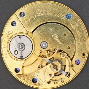 May include: Close-up of a gold-toned pocket watch movement with the inscription "Elgin National Watch Co.  Dexter, St." and "Patented" visible. The watch movement has a intricate design with gears and a balance wheel.