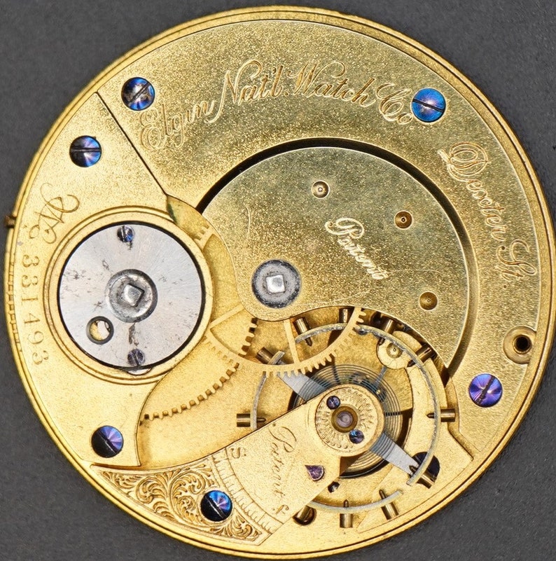 May include: Close-up of a gold-toned pocket watch movement with the inscription "Elgin National Watch Co.  Dexter, St." and "Patented" visible. The watch movement has a intricate design with gears and a balance wheel.