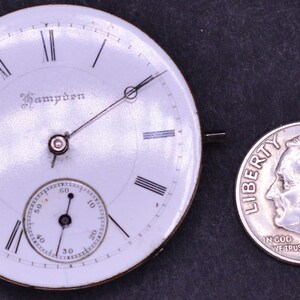 K&ouml;nnte beinhalten: Ein wei&szlig;es Taschenuhrzifferblatt mit schwarzen r&ouml;mischen Ziffern f&uuml;r die Stunden und einem kleineren Hilfszifferblatt f&uuml;r die Sekunden. Das Zifferblatt der Uhr ist mit "Hampden" signiert und etwa so gro&szlig; wie eine deutsche Zehn-Cent-M&uuml;nze.