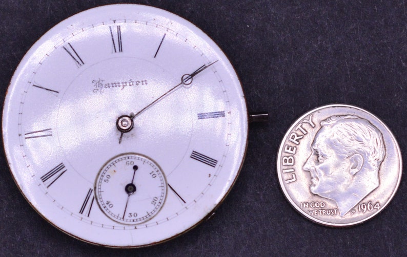 K&ouml;nnte beinhalten: Ein wei&szlig;es Taschenuhrzifferblatt mit schwarzen r&ouml;mischen Ziffern f&uuml;r die Stunden und einem kleineren Hilfszifferblatt f&uuml;r die Sekunden. Das Zifferblatt der Uhr ist mit "Hampden" signiert und etwa so gro&szlig; wie eine deutsche Zehn-Cent-M&uuml;nze.