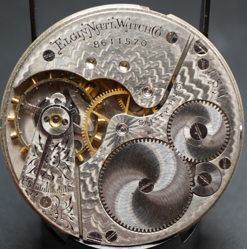 Op de afbeelding: Close-up van een zilveren zakhorloge-uurwerk met ingewikkelde tandwielen en de inscriptie "Elgin Natl. Watch Co. 8611570 Safety Pinion".