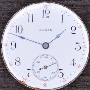 Puede incluir: Una esfera de reloj de bolsillo blanca con marcadores de hora en n&uacute;meros romanos negros y marcadores de minutos en n&uacute;meros ar&aacute;bigos negros. La esfera del reloj tiene un subdial blanco m&aacute;s peque&ntilde;o con marcadores de n&uacute;meros ar&aacute;bigos negros. La esfera del reloj tambi&eacute;n tiene la palabra "ELGIN" impresa en negro.