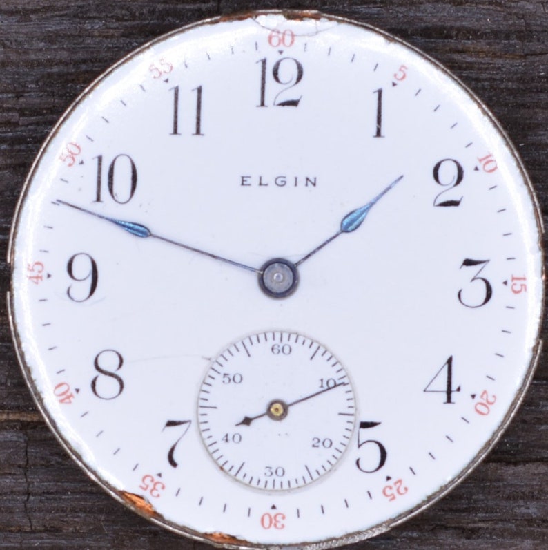 Puede incluir: Una esfera de reloj de bolsillo blanca con marcadores de hora en n&uacute;meros romanos negros y marcadores de minutos en n&uacute;meros ar&aacute;bigos negros. La esfera del reloj tiene un subdial blanco m&aacute;s peque&ntilde;o con marcadores de n&uacute;meros ar&aacute;bigos negros. La esfera del reloj tambi&eacute;n tiene la palabra "ELGIN" impresa en negro.