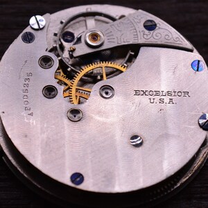 May include: Close-up of a silver pocket watch with a gold-colored gear mechanism. The watch face has the text "EXCELSIOR U.S.A." and the number "A2005235".