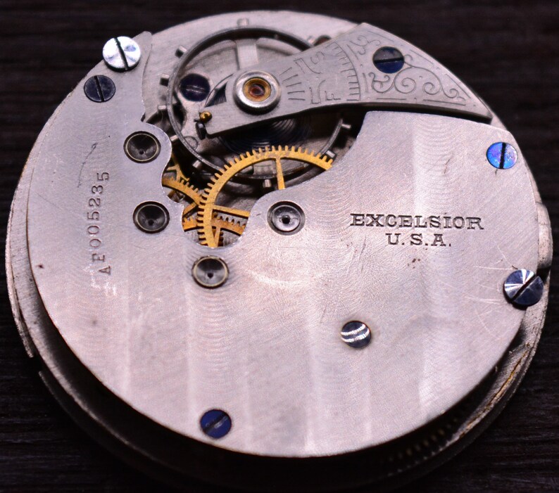 May include: Close-up of a silver pocket watch with a gold-colored gear mechanism. The watch face has the text "EXCELSIOR U.S.A." and the number "A2005235".