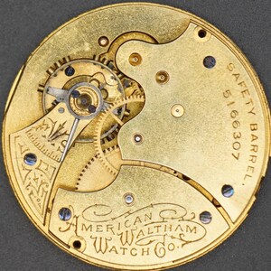 Puede incluir: Primer plano del mecanismo interno de un reloj de bolsillo dorado con el texto "SAFETY BARREL 5166307 AMERICAN WALTHAM WATCH CO." inscrito en la parte posterior.