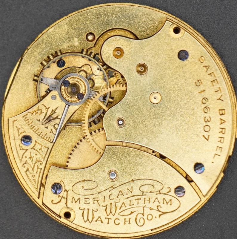 Puede incluir: Primer plano del mecanismo interno de un reloj de bolsillo dorado con el texto "SAFETY BARREL 5166307 AMERICAN WALTHAM WATCH CO." inscrito en la parte posterior.