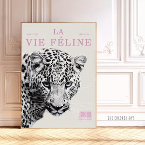 May include: A framed art print featuring a black and white leopard face with a light beige background. The text "LA VIE FÉLINE" is in pink, along with other text and a barcode. The artwork is displayed in a room with white walls and a wooden floor.