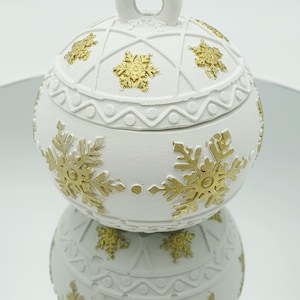May include: A white, round decorative box with a lid, adorned with gold snowflake designs. The box has a loop on top and is reflected in the surface below. The design is reminiscent of a Christmas ornament.