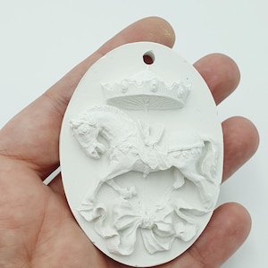 Op de afbeelding: Wit ovaal ornament met een carrousel paard ontwerp. Het paard is afgebeeld met een kroon en een decoratieve strik. Het ornament heeft een gat aan de bovenkant om op te hangen.