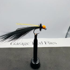 May include: A black and yellow fishing fly with an orange bead head. The fly has a black body with long, flowing fibers and yellow accents. The text "Garage Ted Flies" is visible in the background.