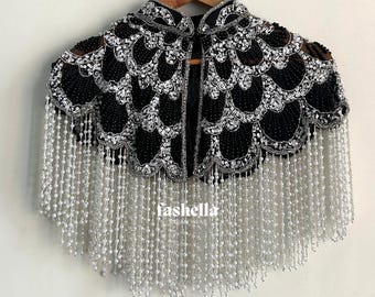 May include: A black and silver beaded cape with a scalloped design and long beaded fringe. The cape is hanging on a wooden hanger against a white wall. The text "fashella" is visible.