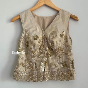 May include: A sleeveless, beige top with a V-neckline and a floral design embroidered with sequins. The top has a scalloped hemline.