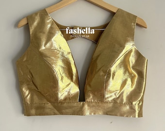May include: A gold sleeveless blouse with a deep V-neckline. The blouse has a shiny, metallic finish and is made of a soft, flowing fabric. The brand name "fashella INDIAN WEAR" is printed on the front of the blouse.