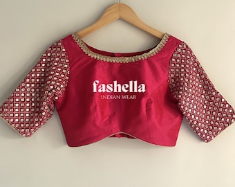 May include: A pink silk blouse with gold trim and a geometric pattern on the sleeves. The blouse has a scoop neckline and short sleeves. The text "fashhella INDIAN WEAR" is printed on the front of the blouse.