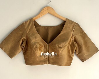 Puede incluir: Una blusa de color dorado con mangas cortas y cuello en V. La blusa está colgada de una percha de madera sobre un fondo blanco. El texto "fashella INDIAN WEAR" está impreso en la parte delantera de la blusa.