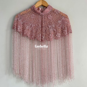 May include: A pink beaded cape with a high collar and fringe detail. The cape is embellished with intricate beadwork and hangs from a wooden hanger. The text "fashella" is visible on the cape.