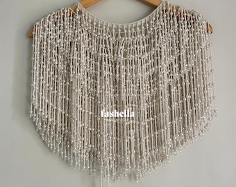 May include: A cream-colored beaded cape hangs on a wooden hanger. The cape is made of rows of small, round beads, creating a fringed effect at the bottom. The word "fashella" is printed on the cape.