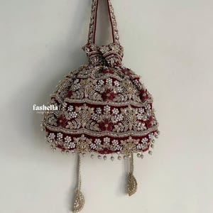 May include: A deep red velvet drawstring pouch bag featuring intricate gold embroidery, white pearl bead accents, and floral motifs. The bag includes a matching embroidered handle and two dangling tassels with decorative gold and pearl embellishments.