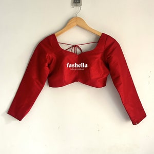 May include: A red long-sleeved blouse with a square neckline and a tie-up back detail. The blouse is made of a smooth, silky fabric and has a cropped fit. The brand name "f'ashella INDIAN WEAR" is printed on the front of the blouse.