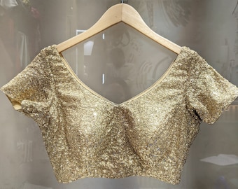 May include: A short-sleeved blouse covered in small, reflective gold sequins. The garment features a V-neckline, structured seams across the front bodice, and is displayed on a light-colored wooden hanger against a neutral, semi-transparent background.