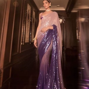 May include: A glamorous lavender and purple sequined saree. The fabric transitions from a light lavender to a deep purple at the border. The saree is draped to show off its shimmering texture and elegant design, perfect for special occasions.