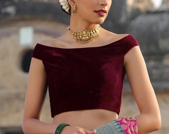 May include: A woman wearing a maroon velvet off-the-shoulder top and gold jewellery, including a necklace and earrings. She is also wearing a pink and blue patterned sari. The background is a blurred outdoor scene.