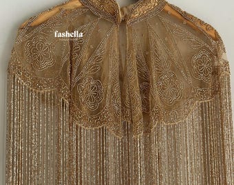 Puede incluir: Una capa dorada con intrincados abalorios y flecos. La capa presenta un cuello alto y un dobladillo festoneado, con diseños florales y de hojas. El nombre de la marca "fashella" es visible en el lado izquierdo.