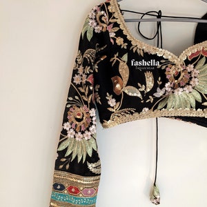 May include: Black crop top with long sleeves featuring floral embroidery and sequins. The top has a tie closure at the back and a gold trim around the neckline. The sleeves have a multi-colored trim with sequins.
