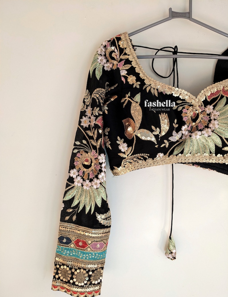 May include: Black crop top with long sleeves featuring floral embroidery and sequins. The top has a tie closure at the back and a gold trim around the neckline. The sleeves have a multi-colored trim with sequins.