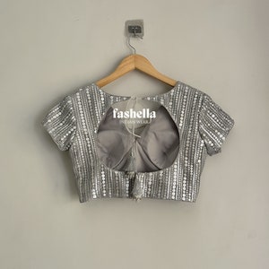 May include: A silver sequined blouse with a keyhole back and a tassel detail. The blouse is hanging on a wooden hanger against a white wall. The text "fashella INDIAN WEAR" is visible on the blouse.