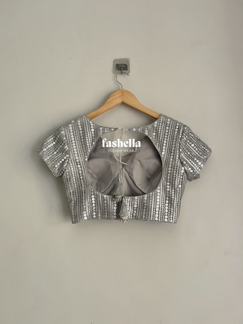 May include: A silver sequined blouse with a keyhole back and a tassel detail. The blouse is hanging on a wooden hanger against a white wall. The text "fashella INDIAN WEAR" is visible on the blouse.