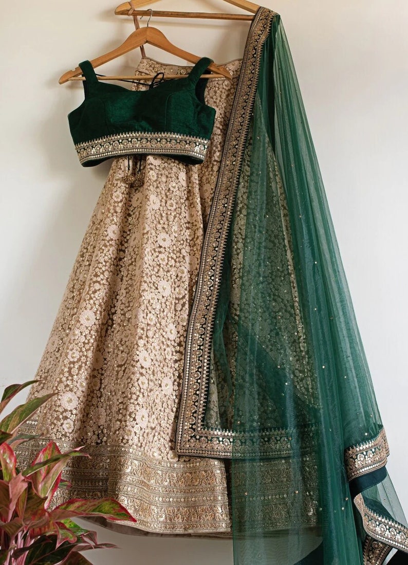 May include: A beige embroidered lehenga set with a dark green velvet top and matching dupatta. The lehenga features intricate floral patterns and gold embellishments. The dupatta has a gold border and sequin details. The outfit is displayed on a wooden hanger.