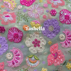 May include: Light green fabric featuring embroidered floral patterns in shades of pink, purple, yellow, and orange. The flowers are adorned with sequins and small mirrors. The fabric has the text "fashella INDIAN WEAR".