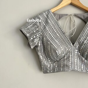 May include: A gray sequined crop top with a V-neckline and short sleeves. The top has a tie-back closure and is decorated with silver sequins.