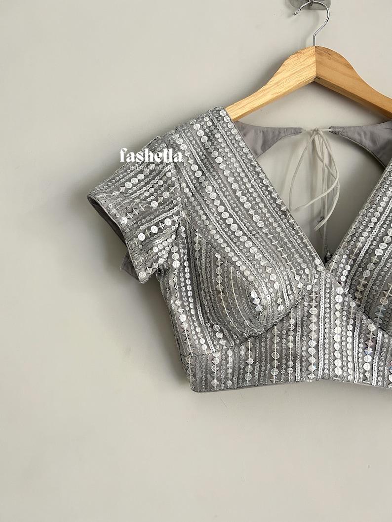 May include: A gray sequined crop top with a V-neckline and short sleeves. The top has a tie-back closure and is decorated with silver sequins.