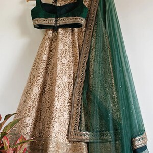 May include: A traditional Indian outfit in gold and green. The skirt is heavily embroidered in gold, and the top is a deep green. A matching green scarf with gold detailing is draped over the outfit. The outfit is displayed on a wooden hanger.