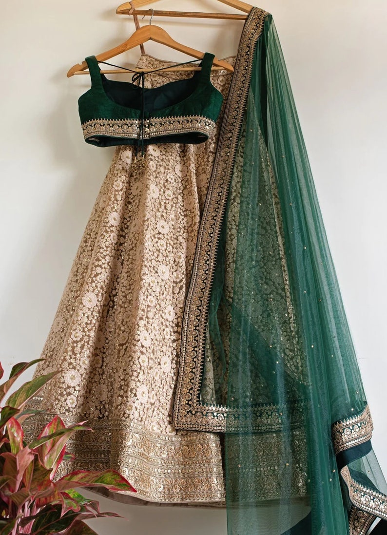 May include: A traditional Indian outfit in gold and green. The skirt is heavily embroidered in gold, and the top is a deep green. A matching green scarf with gold detailing is draped over the outfit. The outfit is displayed on a wooden hanger.