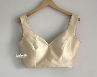 May include: A sleeveless, cream-colored blouse with a deep V-neckline and wide shoulder straps, hanging on a wooden hanger. The fabric has a subtle sheen. The text 'fashella INDIAN WEAR' is printed in white on the lower left side of the garment.