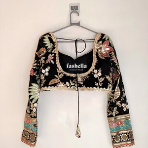 May include: A black embroidered blouse with long sleeves and a tie closure at the neckline. The blouse features floral and paisley patterns in gold, pink, green, and blue thread. The brand name "fashella INDIAN WEAR" is printed on the front of the blouse.