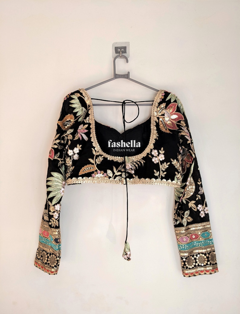 May include: A black embroidered blouse with long sleeves and a tie closure at the neckline. The blouse features floral and paisley patterns in gold, pink, green, and blue thread. The brand name "fashella INDIAN WEAR" is printed on the front of the blouse.