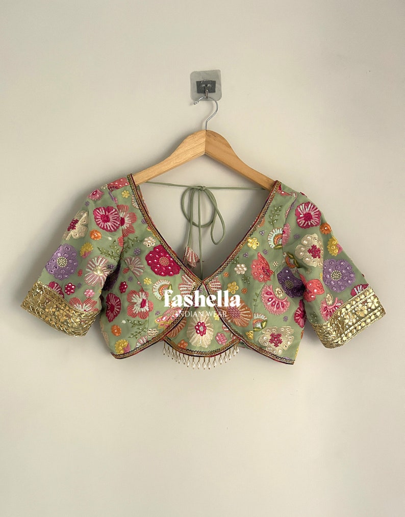 May include: A light green embroidered blouse with a deep V-neck and short sleeves. The blouse features colourful floral embroidery, gold trim on the sleeves, and a tie closure at the back. The brand name "fashella" is visible.