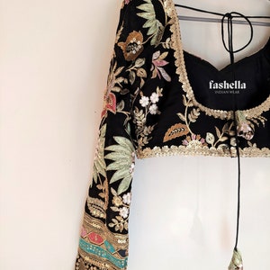 May include: A black embroidered blouse with long sleeves and a tie closure at the back. The blouse features floral and leaf patterns in gold, green, pink, and white thread. The sleeves have a gold and turquoise trim. The blouse is labeled "fashhella INDIAN WEAR".