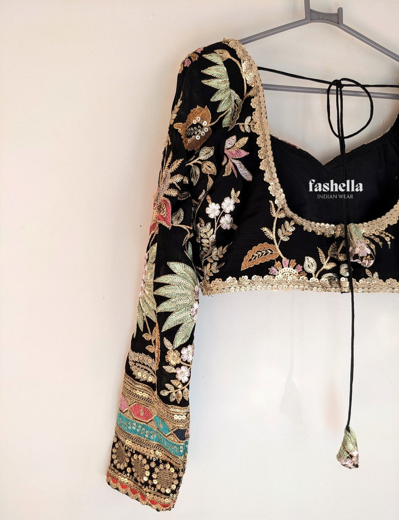 May include: A black embroidered blouse with long sleeves and a tie closure at the back. The blouse features floral and leaf patterns in gold, green, pink, and white thread. The sleeves have a gold and turquoise trim. The blouse is labeled "fashhella INDIAN WEAR".